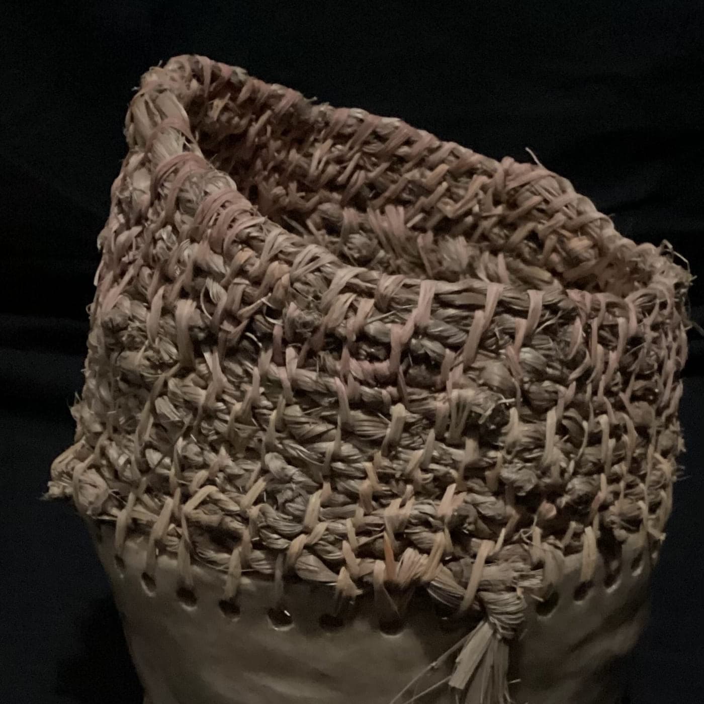 Tall coiled fibre vessel over a burnished clay base, photographed against black