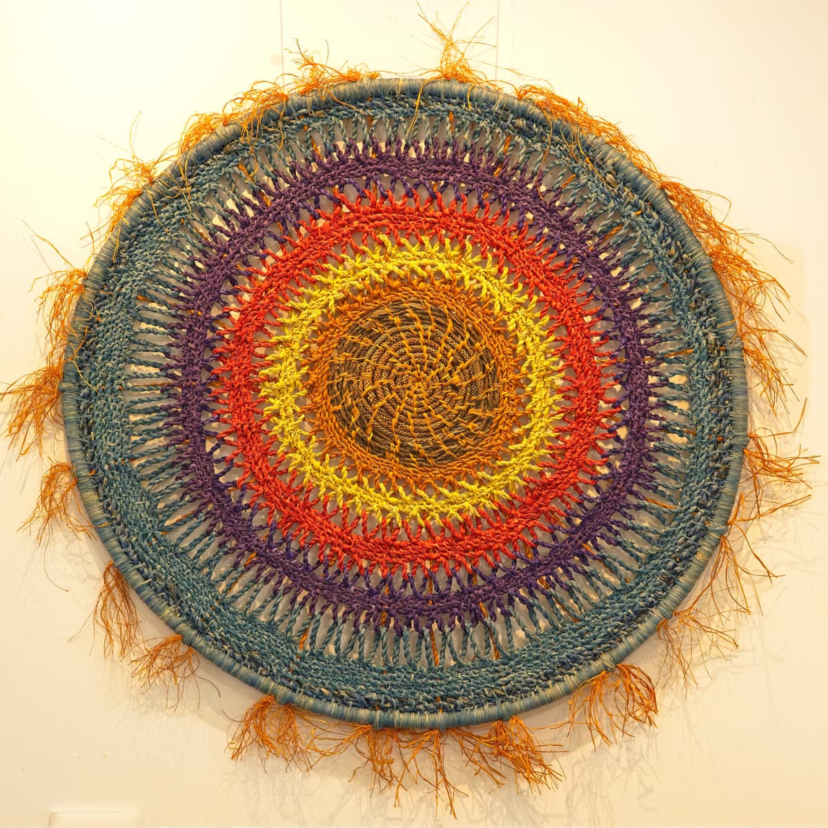 A large circular coiled mandala in red, gold and teal with a fringed edge