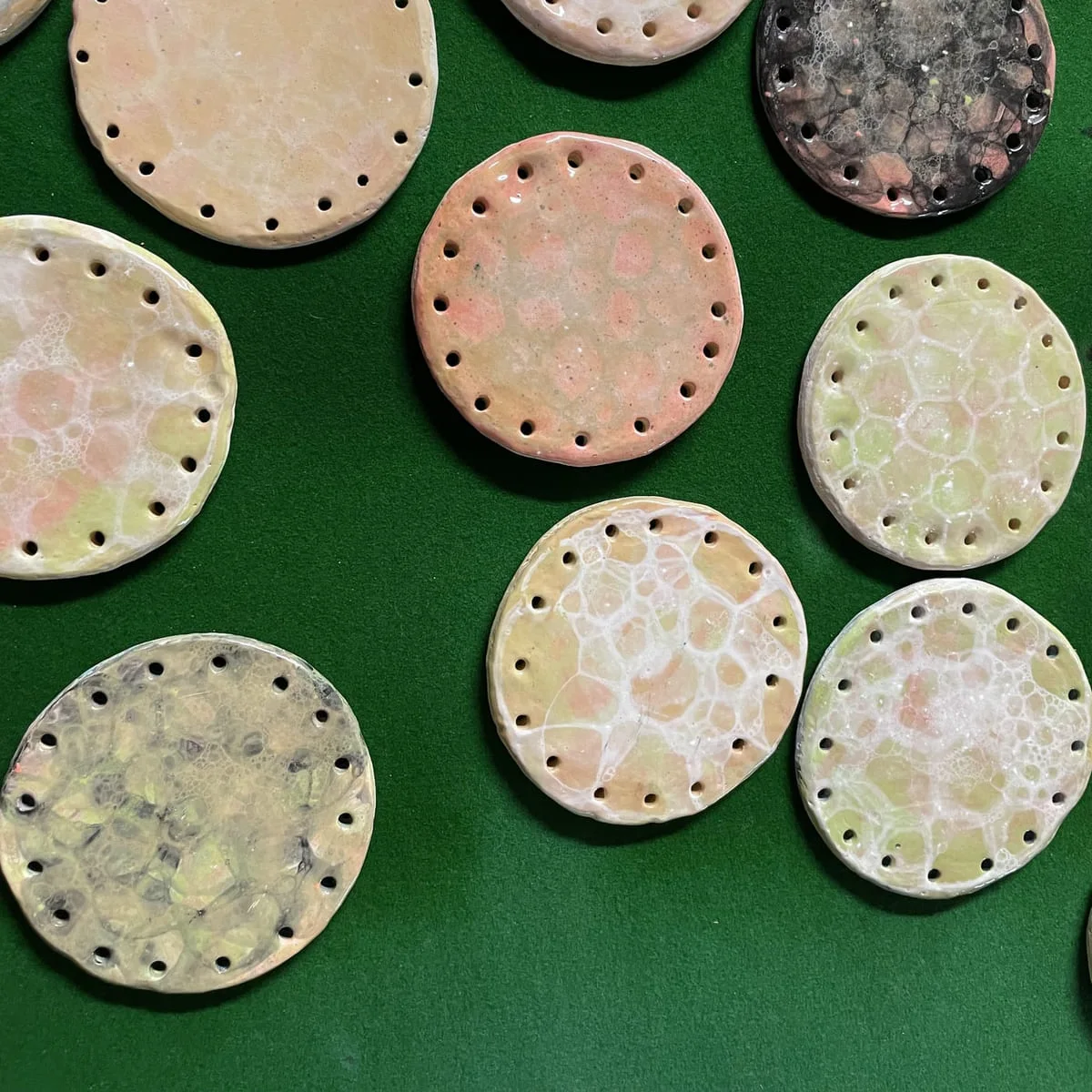 Eight round clay discs with pierced edges and soft pastel glazes on green baize