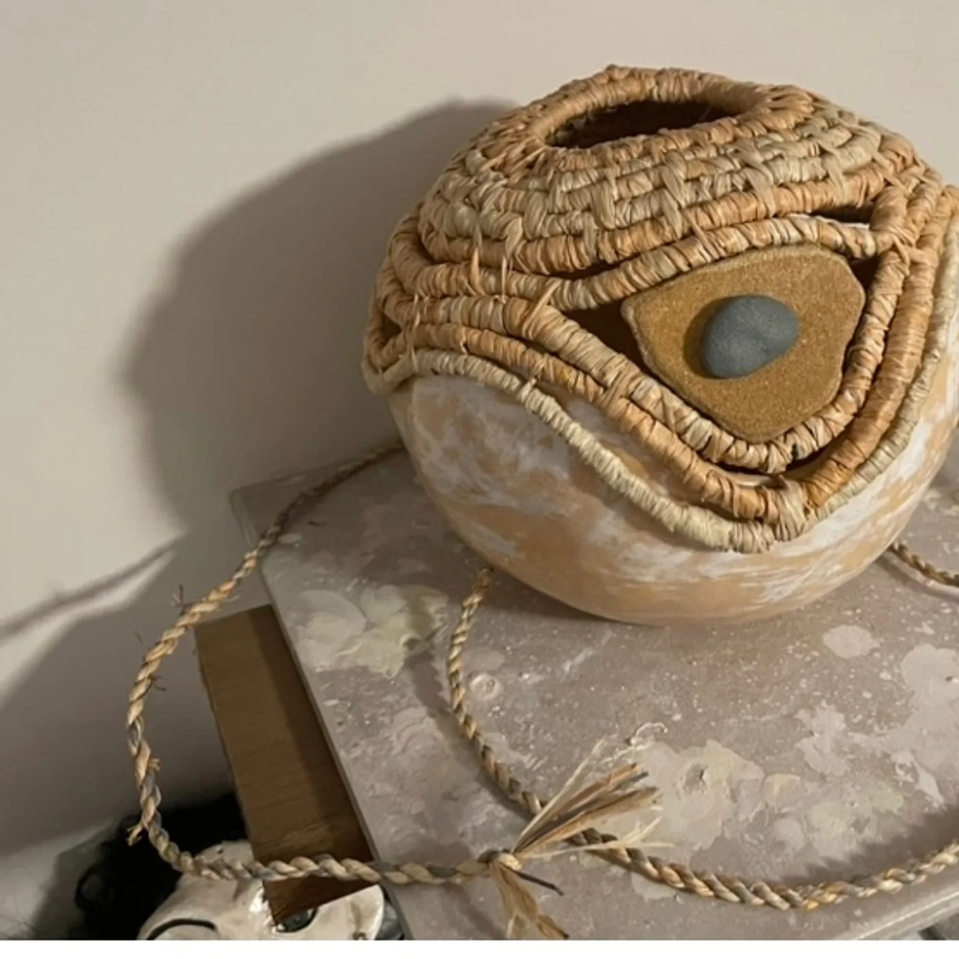 A pale clay sphere wrapped in coiled natural fibre around a set river stone, on a speckled plinth