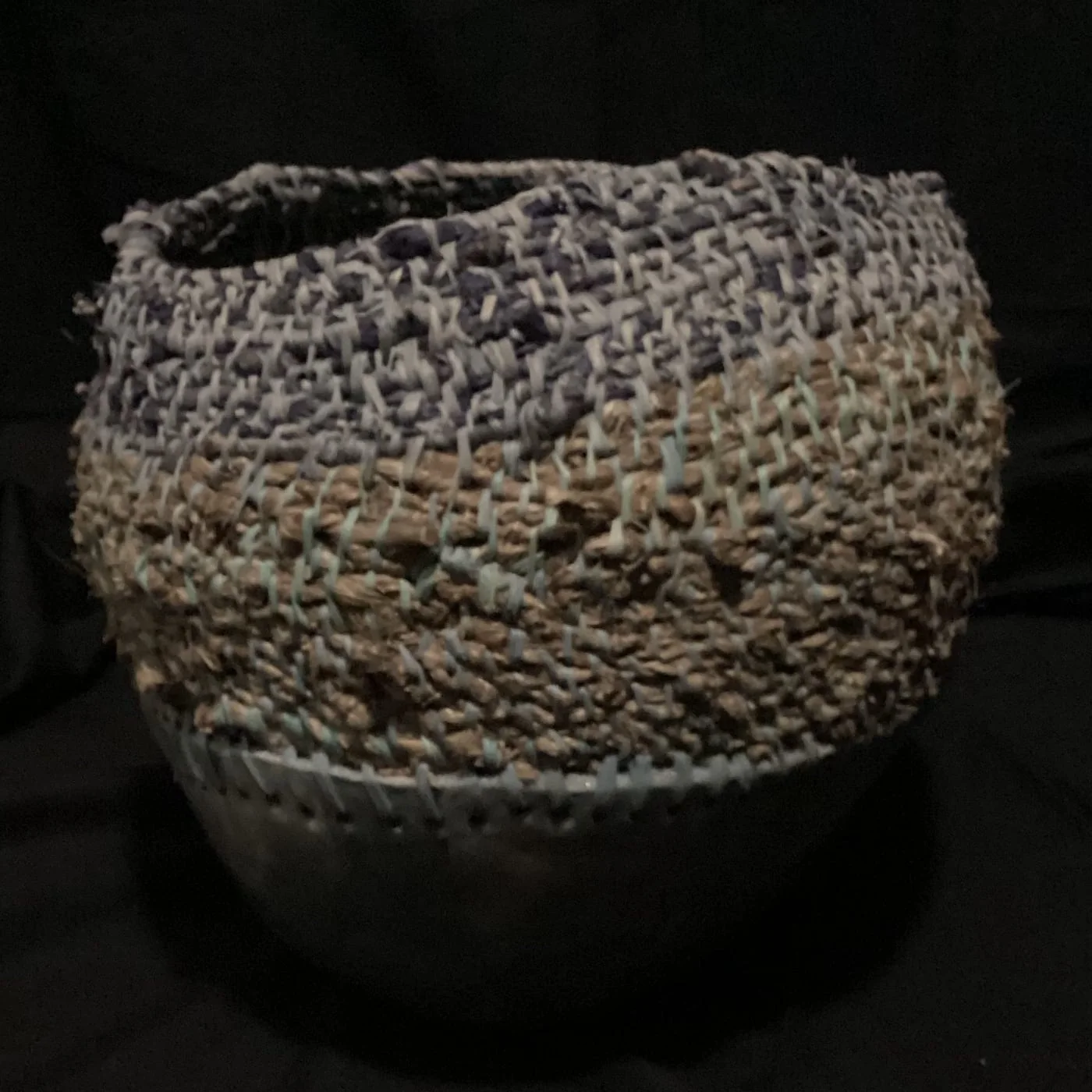 An open, loosely coiled fibre vessel over a pale clay base, photographed against black