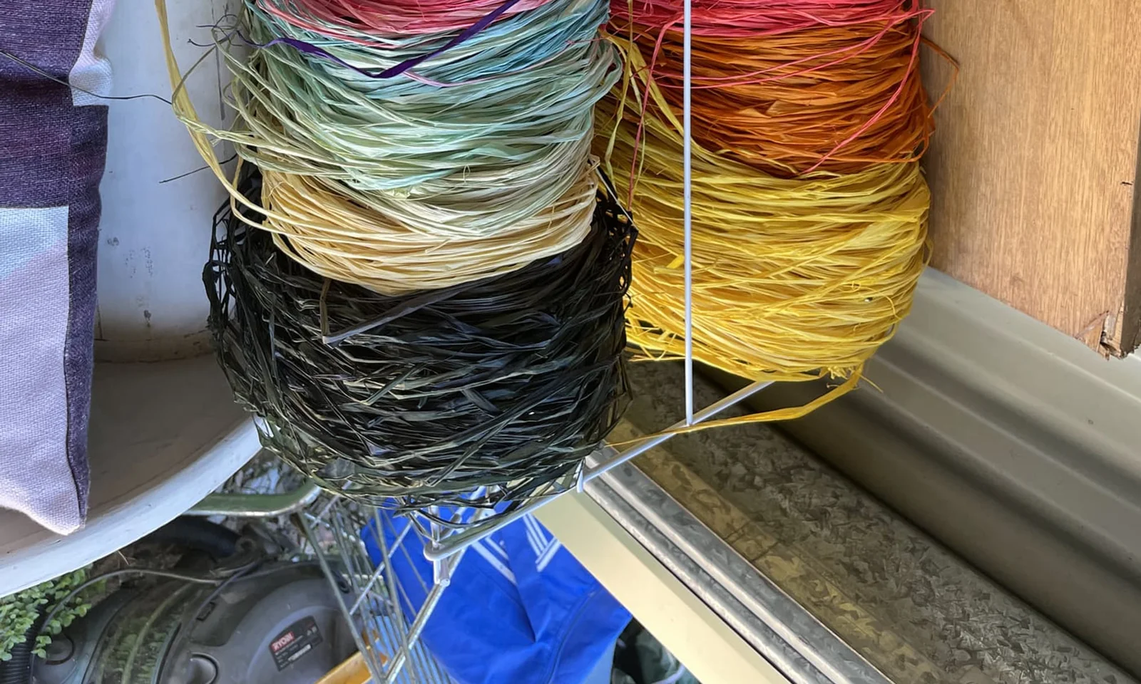 Bundles of dyed raffia in green, yellow, orange, pink and black stacked on a shelf by a window