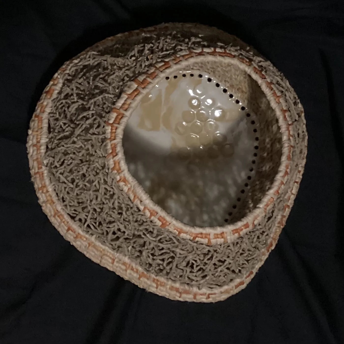 An oval vessel with a fine coiled fibre wall and a pierced glazed clay base inside, against black