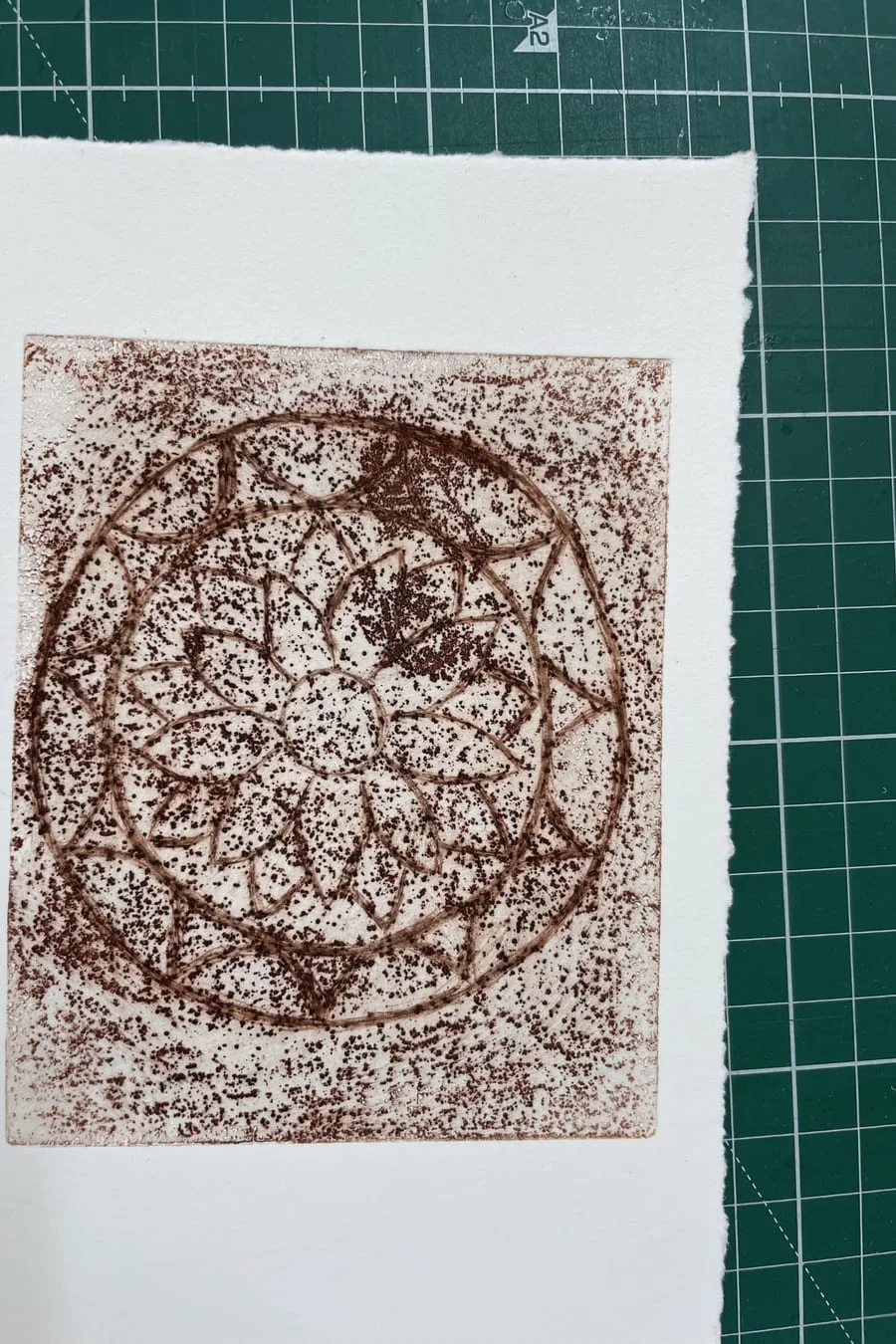 A circular brown collagraph print on deckle-edged paper, on a cutting mat