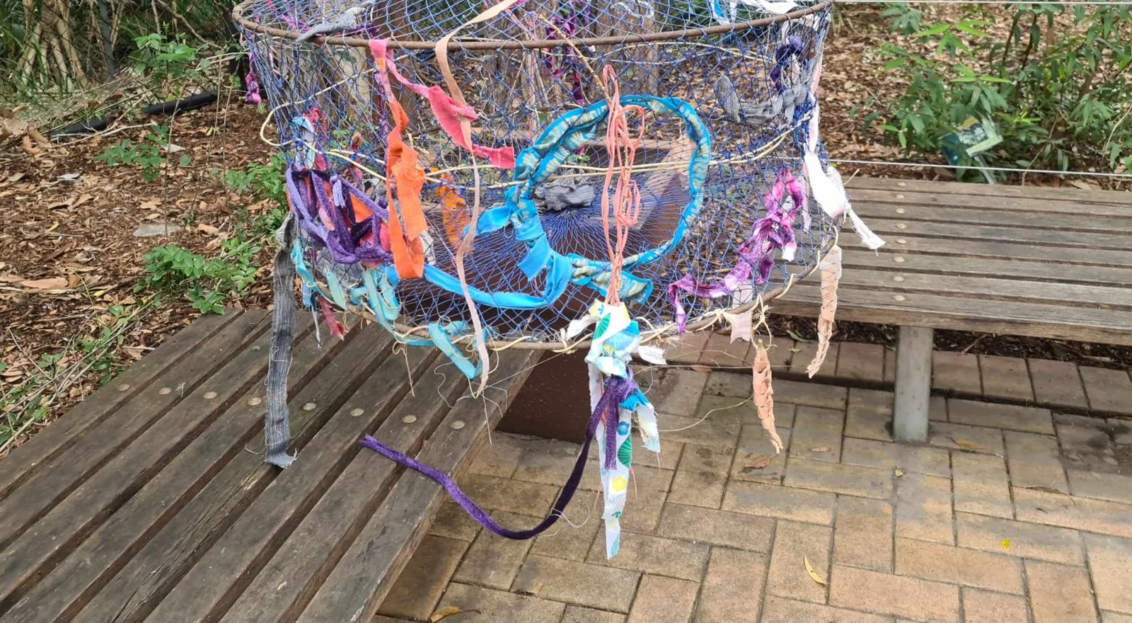 A hanging canopy of netting threaded with coloured ribbon and fibre above a garden bench