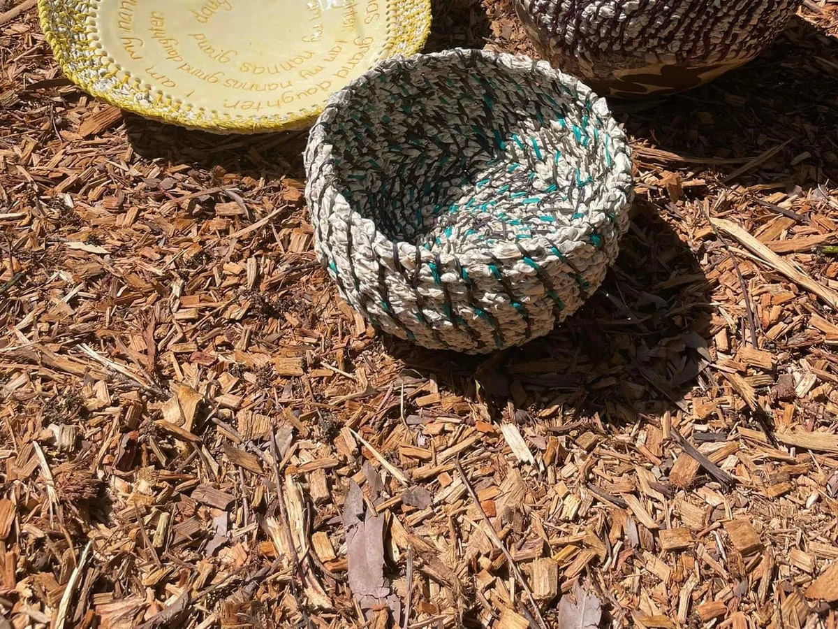 A small coiled bowl in teal and cream on bark mulch, beside a yellow plate