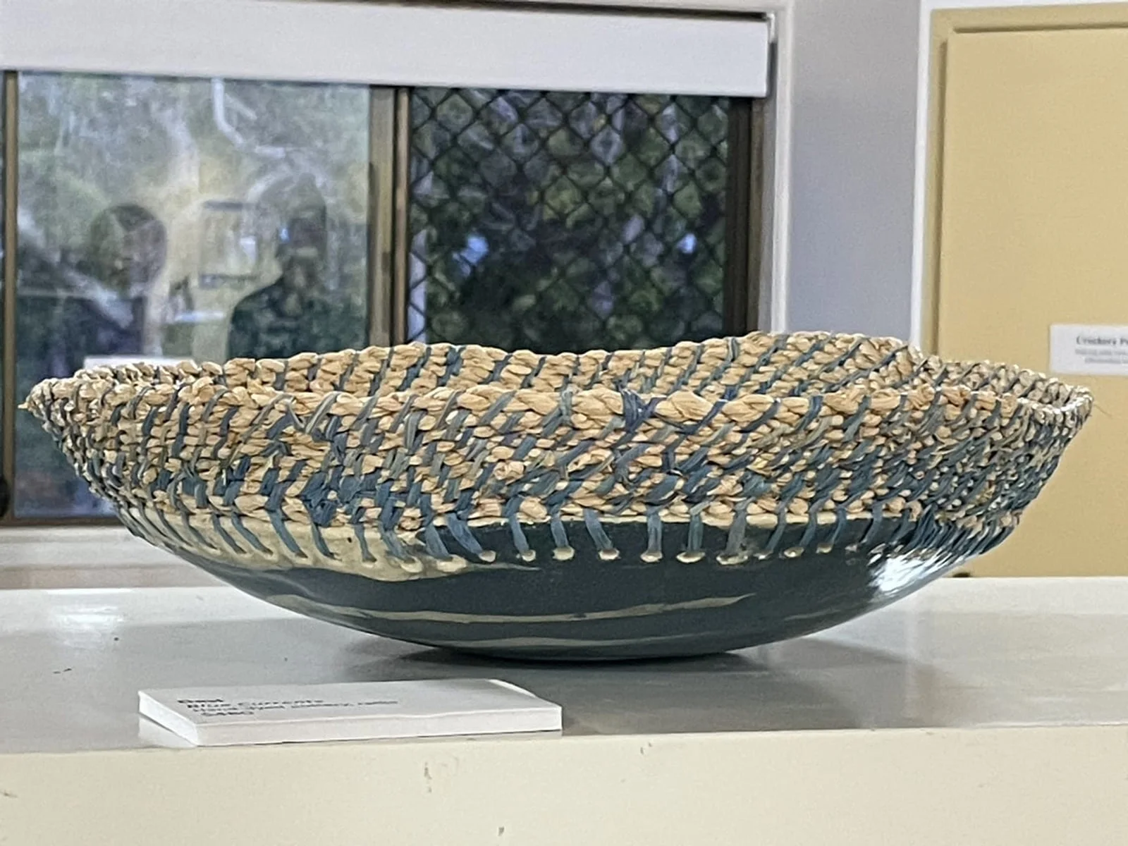 A wide, shallow coiled bowl with a blue clay base on a white gallery shelf